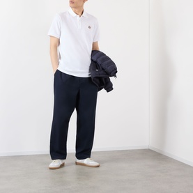 Model wearing Moncler white cotton polo shirt with navy trousers and white trainers, holding a navy jacket