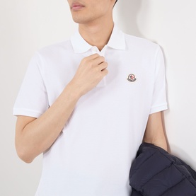Model wearing Moncler white cotton polo shirt with logo badge, styled with navy jacket over arm.