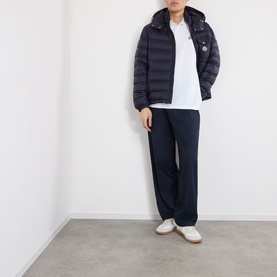 Model wearing Moncler white cotton polo, styled with navy quilted hooded jacket, navy trousers and white trainers.