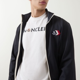 Model wearing Moncler natural cotton T-shirt with logo print, styled with a navy zip-up hoodie featuring tricolour badge detail.