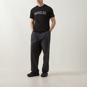 Model wearing Moncler black cotton T-shirt with logo print, styled with dark grey trousers and black trainers.