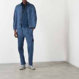 Model wearing blue Stone Island overshirt jacket with matching blue joggers, layered over navy zip-up, styled with grey trainers