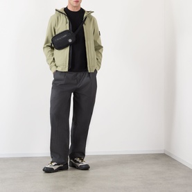 Model wearing Stone Island Softshell-R_e.dye&reg; Wind Resistant Jacket in Laurel Green with black tee, grey trousers and chunky trainers, black belt bag