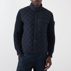 Model wearing Stone Island blue quilted gilet with snap-button fastening, badge patch on sleeve, styled over navy knit sleeves