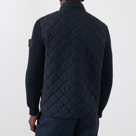 Model wearing Stone Island blue quilted casual jacket, back view, with badge patch on sleeve, styled over navy long-sleeve top