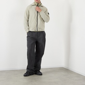 Model wearing Stone Island Compass Logo Ultralight Windbreaker in sand, styled with dark charcoal trousers and black shoes.
