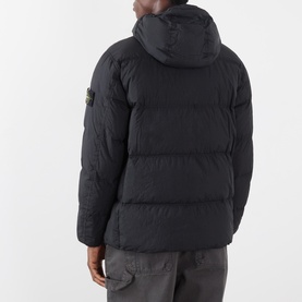 Model wearing Stone Island black hooded puffer jacket, rear view showing compass badge on left sleeve, styled with grey trousers.