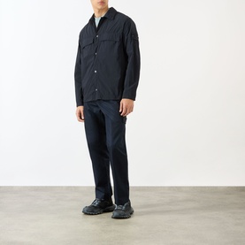 Model wearing Stone Island blue polyamide casual shirt with chest pockets, styled with navy trousers and black trainers.