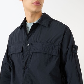 Model wearing Stone Island blue polyamide casual shirt with chest pockets, button placket, and signature sleeve badge, styled over light blue crew