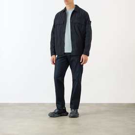 Model wearing Stone Island blue polyamide casual shirt over light grey top, styled with dark navy trousers and black trainers.