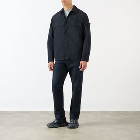 Model wearing Stone Island Tonal Logo Badge Zip Overshirt in blue, styled with dark trousers and grey trainers.