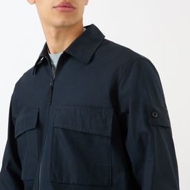 Model wearing Stone Island Tonal Logo Badge Zip Overshirt in blue, with chest flap pockets and sleeve logo badge visible.