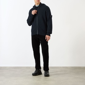 Model wearing Stone Island Logo Badge Corduroy Trousers in black, styled with a dark zip-up hooded jacket and black boots.