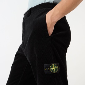 Model wearing Stone Island Logo Badge Corduroy Trousers in black, close-up showing logo badge patch and hand pocket detail.