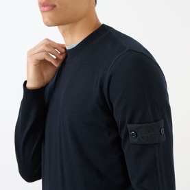 Model wearing Stone Island blue virgin wool crew neck knit with signature sleeve badge detail, layered over light blue top.