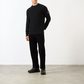 Model wearing black crew neck sweatshirt with Stone Island arm badge, styled with black trousers and black trainers.