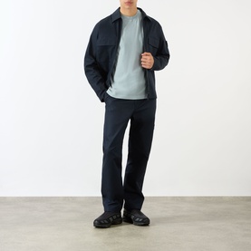 Model wearing Stone Island blue cotton chino trousers, styled with a navy zip jacket and sage green T-shirt, black sandals.