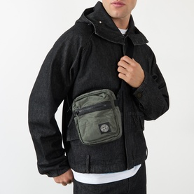 Model carrying Stone Island Logo Badge Crossbody Bag in green, with front zip pocket and logo badge, worn over black jacket