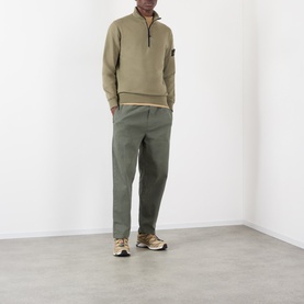 Model wearing Stone Island Military Green cotton half-zip sweatshirt with arm badge, styled with khaki trousers and beige trainers.