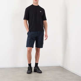 Model wearing Stone Island Compass Logo Bermuda Shorts in navy blue with cargo pockets, styled with black t-shirt and chunky black trainers.