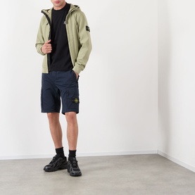Model wearing Stone Island Compass Logo Bermuda Shorts in navy blue with cargo pockets, paired with sage green zip-up jacket and black trainers.