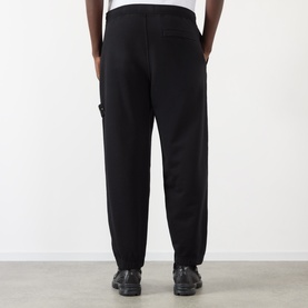 Model wearing Stone Island black cotton sweatpants, rear view, with utility-style seam detailing, styled with dark lace-up shoes.