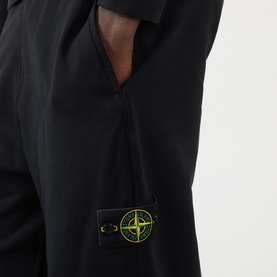 Model wearing Stone Island black cotton sweatpants, close-up of signature compass badge patch on thigh and side pocket detail.