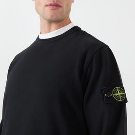 Model wearing Stone Island black cotton sweatshirt with signature compass badge patch on left sleeve, styled with white crew-neck underneath.