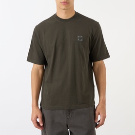Model wearing Stone Island Compass Logo T-Shirt in coal grey, crew neck with small logo patch, styled with grey trousers.