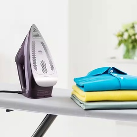 Braun TexStyle 7 Pro Steam Iron in purple and white, upright on ironing board beside folded colourful shirts