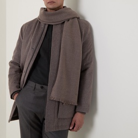 Model wearing a taupe woven scarf with fringe edges, styled with a matching coat and dark trousers