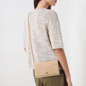 Model carrying a small beige crossbody bag with gold chain strap and metal logo detail, styled with a cream knit top and khaki trousers