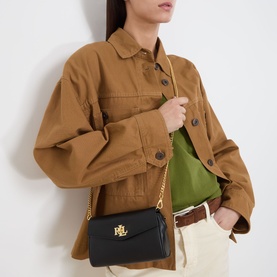 Model carrying small black crossbody bag with gold chain strap and logo hardware, styled with tan jacket and cream trousers