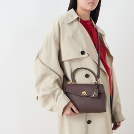 Model carrying Lauren Farrah Small Satchel Bag in chestnut brown leather, gold-tone hardware, top handle, worn over beige trench coat.