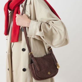 Model carrying Lauren Marcy Charm Crossbody Bag in chestnut brown suede leather with gold-tone hardware and branded charm, styled with beige trench