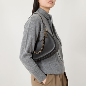 Model carrying a black crescent shoulder bag with gold chain strap, styled with a grey knit jacket and tan trousers
