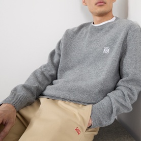 Model wearing Loewe Anagram Logo Sweater in grey melange, crew neck wool knit with chest logo embroidery, styled with beige trousers.