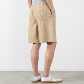Model wearing Loewe beige cotton shorts, back view showing embroidered pocket detail and elasticated waist, styled with white trainers.