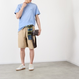 Model wearing Loewe beige cotton shorts with a light blue T-shirt and white trainers, holding a patchwork bag.