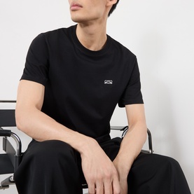 Model wearing Loewe black cotton T-shirt with small chest logo, paired with black trousers, seated pose.