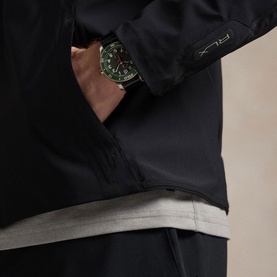 Model wearing RLX Golf black casual jacket, hands in pockets, styled with grey layered top and green-dial watch visible at cuff.