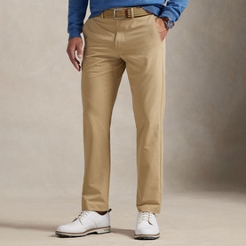 Model wearing RLX Golf beige tailored golf trousers with brown belt, styled with white leather golf shoes and blue polo shirt.