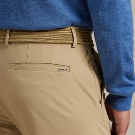 Model wearing RLX Golf beige trousers, close-up of back pocket with logo detail, styled with a woven tan belt and blue knit top.
