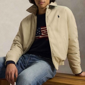 Model wearing Ralph Lauren Khaki Uniform casual jacket over navy American flag knit jumper and blue jeans, seated.