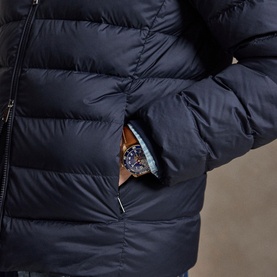 Model wearing navy quilted puffer jacket, hand in zip pocket revealing a blue-dial watch with brown leather strap