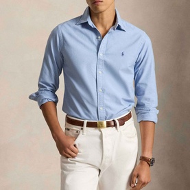 Model wearing Ralph Lauren Pony Logo Slim Fit Oxford Shirt in Chambray Blue, styled with white trousers and a brown leather belt.