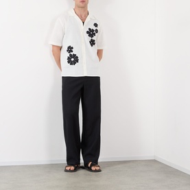 Model wearing Wax London Didcot Floral Shirt in ecru with black floral embroidery, camp collar, styled with black wide-leg trousers and sandals