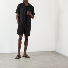 Model wearing Wax London Didcot Linen Bowling Shirt in black, styled with matching black shorts and dark leather sandals.
