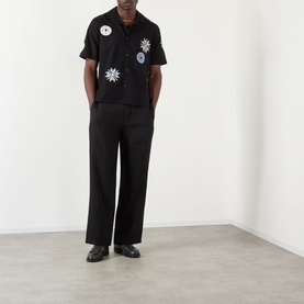 Model wearing Wax London Ditcot Embellished Bowling Shirt in black with floral patches, styled with wide-leg black trousers and black leather shoes.