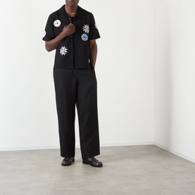 Model wearing Wax London Ditcot Embellished Bowling Shirt in black with floral patches, styled with wide-leg black trousers and black loafers.